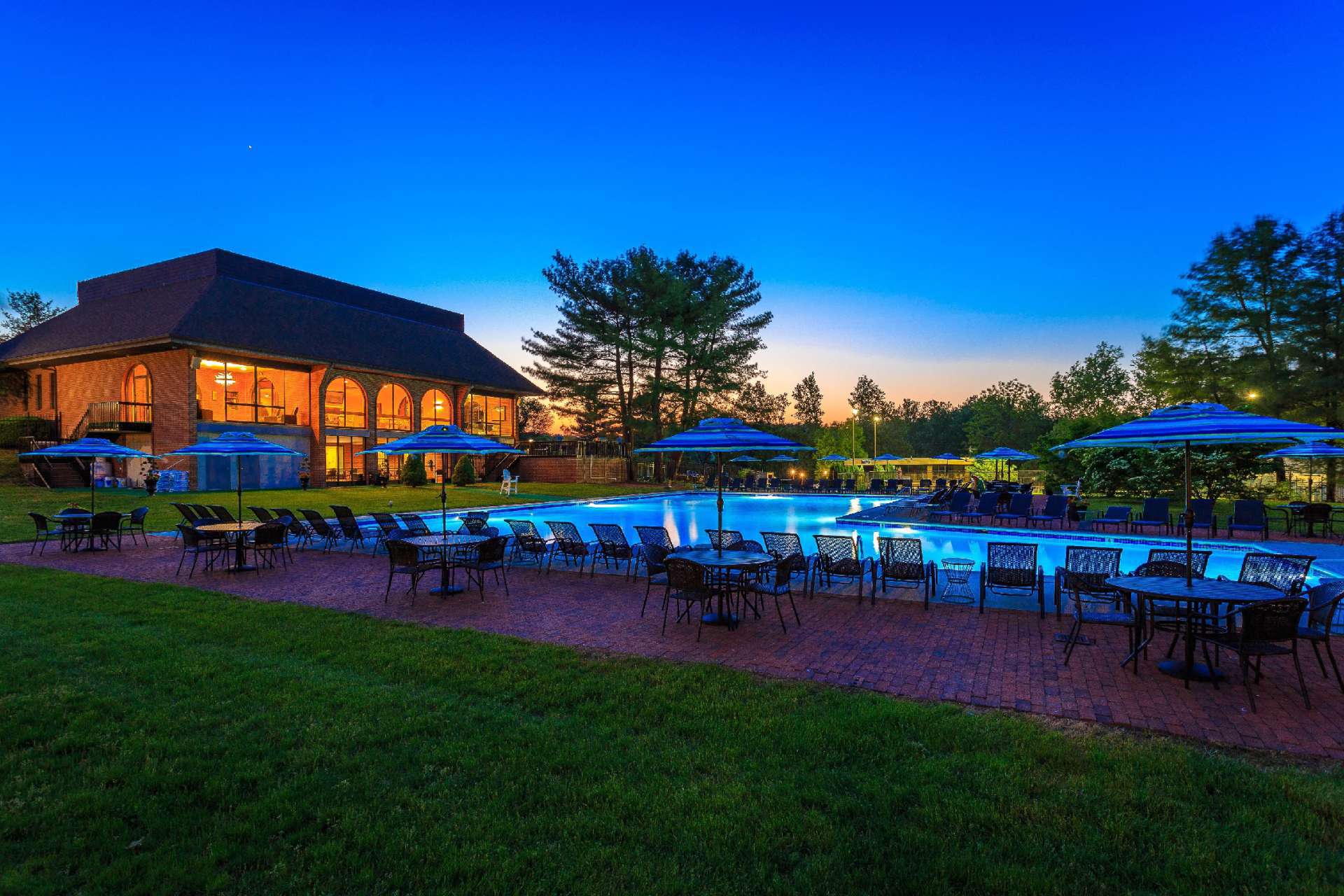 In-ground L shaped pool surrounded with brick patios. Variety of tables, umbrellas, & chairs.