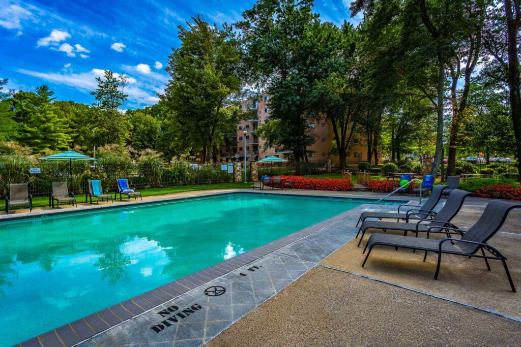 Outdoor swimming pool with lounge chairs, surrounded by greenery and a brick building.